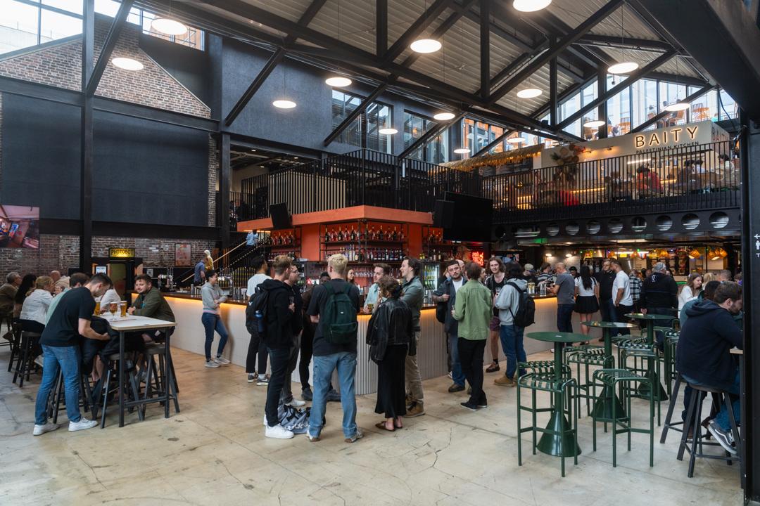 Interior of a modern food hall with high ceilings, exposed beams, and industrial-style lighting. The space is busy with groups of people standing and sitting at tables, socializing and enjoying drinks. At the center is a large bar with an orange canopy, surrounded by bar stools. In the background, there are multiple food stalls and a mezzanine level with additional seating. A sign reading “BAITY” is visible on the upper level.