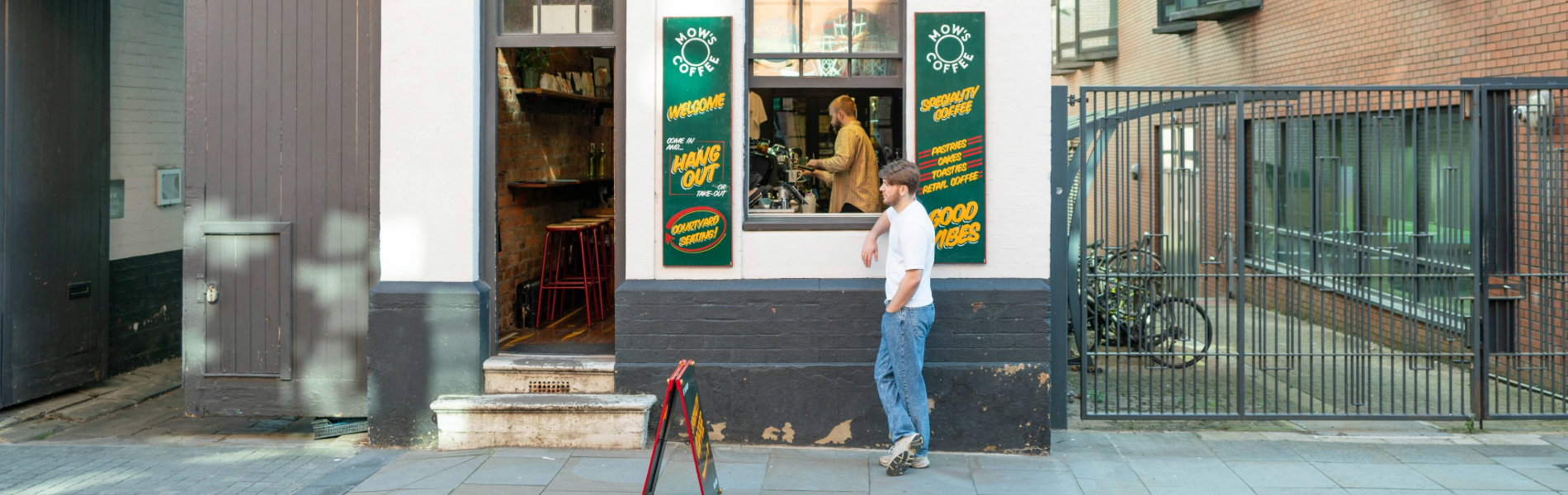 Street view of a café with a white exterior and dark trim, featuring two windows and an open doorway. Green signs with yellow text advertise coffee and good vibes. Inside, a rustic interior with wooden stools and shelves is visible, and a person is preparing drinks at the counter. Outside, another person stands near a sandwich board on the pavement. A black metal gate and bicycles are seen to the right, adding to the urban setting.