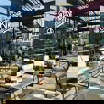 An outdoor terrace with marble-topped tables and green woven chairs, set against a backdrop of modern buildings on a bright, sunny day.