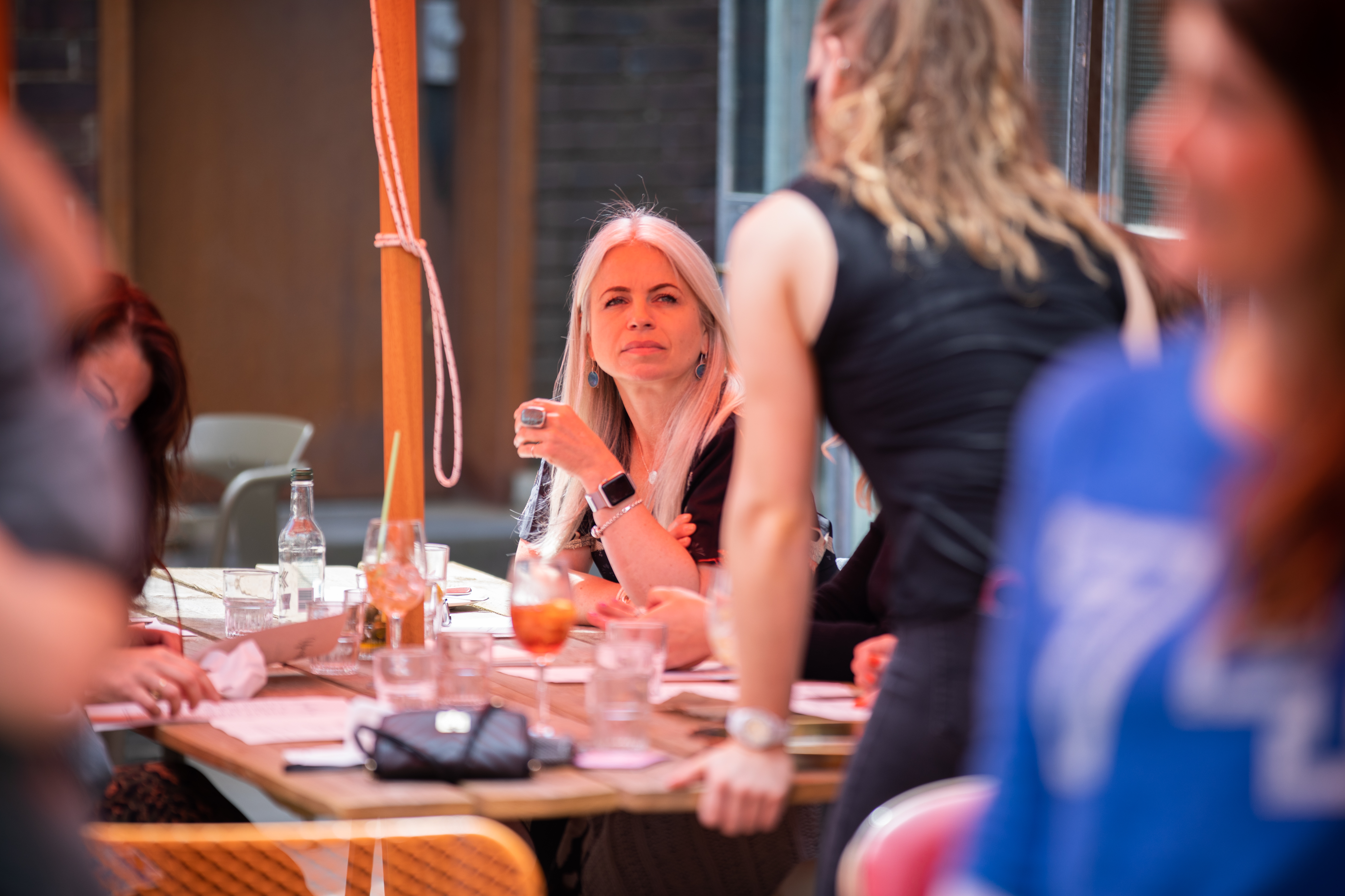 Group of people seated around a wooden table at an indoor venue, engaged in conversation. The table is covered with menus, glasses, and drinks, including a cocktail and a bottle of water. A person in the foreground is standing and leaning toward the table, while others are seated. The setting has warm lighting and a casual atmosphere.