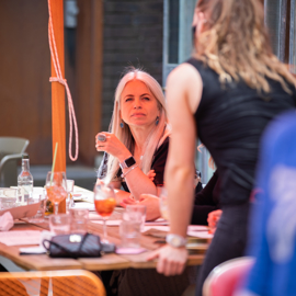 Group of people seated around a wooden table at an indoor venue, engaged in conversation. The table is covered with menus, glasses, and drinks, including a cocktail and a bottle of water. A person in the foreground is standing and leaning toward the table, while others are seated. The setting has warm lighting and a casual atmosphere.