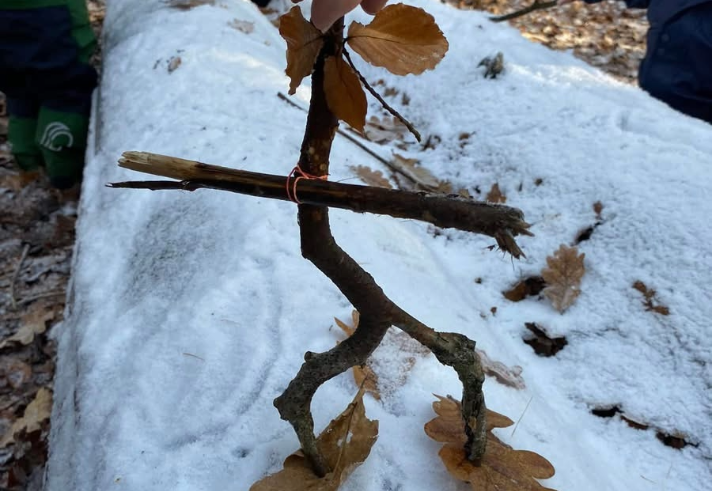 A hand holds up a small figure made from twigs and dried leaves, shaped to resemble a person, standing on snow. The figure has leaf “feet” and “hands”, with a stick for arms and a bent twig for legs. Children in winter clothing are visible in the background, playing in a snowy woodland setting.