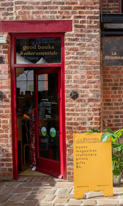 Exterior of a brick building with a red-framed door and windows. The entrance has a sign above reading “good books & other essentials,” and a hanging sign says “La Biblioteka.” A bright yellow board outside lists items such as books, magazines, stationery, prints, and gifts. A green potted plant sits beside the board on a cobbled walkway, creating a welcoming and vibrant storefront.