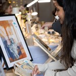 A person views a framed abstract artwork at an art or craft fair. The artwork features warm tones with blue accents. Surrounding the display are books, decorative items, and string lights, creating a cozy and creative atmosphere.
