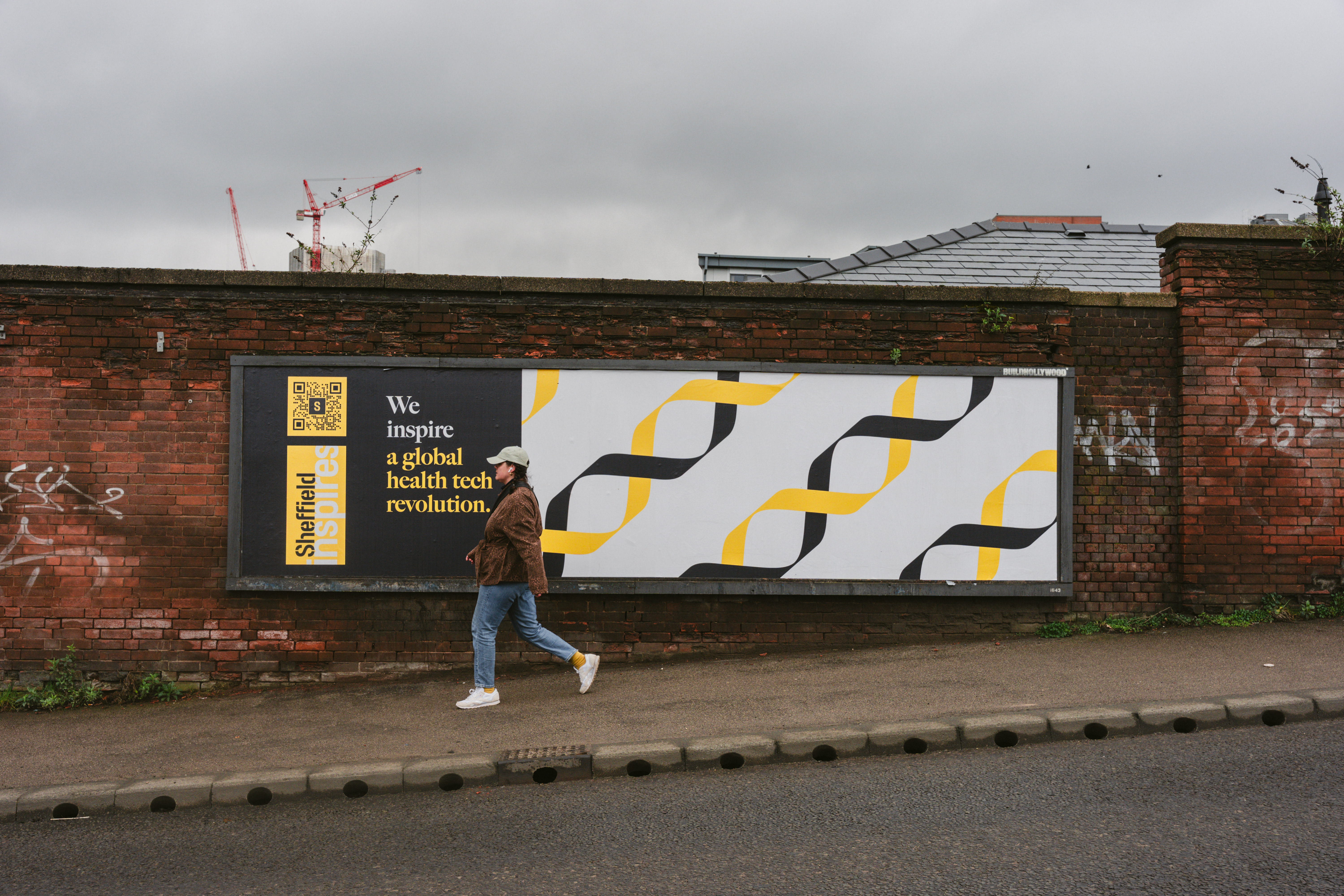 A person walks along a pavement in front of a large brick wall featuring a billboard. The billboard has a modern design with black and yellow geometric DNA-like patterns on a white background. On the left side, there is a black panel with yellow text that reads: “Sheffield” and “We inspire a global health tech revolution.” The sky is overcast, and there are construction cranes visible in the distance above the wall.