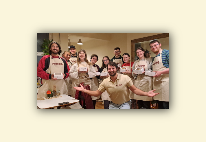 Group of people standing indoors wearing beige aprons with “PARADISO” printed on them, holding white pastry boxes. A person in front kneels with arms open, wearing a matching beige shirt with the same logo. The setting includes wooden tables, chairs, a small flower arrangement, and warm lighting, suggesting a baking or cooking class environment.