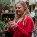 A person holds a small house plant, smiling, in the middle of a plant stall at an indoor market.