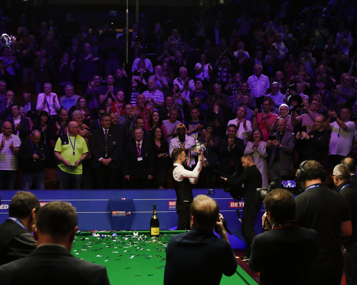 Snooker championship celebration at an indoor arena. A player stands at the snooker table holding a trophy while photographers capture the moment. Green table surface is covered with confetti and a champagne bottle is placed on it. Behind the table, a large crowd of spectators applauds under bright stage lighting.
