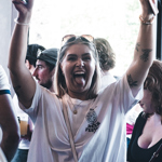 A person in a busy indoor venue raising both arms in the air while holding a phone, with a group of people socialising behind them.