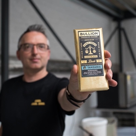 Person holding a gold-wrapped chocolate bar labeled “Bullion Chocolate Maker” with details including “Single Origin Dark Chocolate” and “Tanzania, Morogoro.” The background shows an industrial-style setting with metal beams, white brick walls, and chocolate-making equipment.