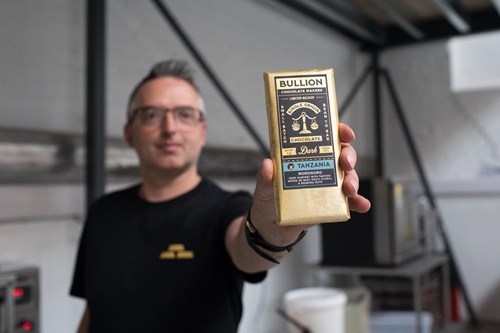 Close-up of a person holding a Bullion chocolate bar labeled 'Dark 70% Tanzania' with packaging featuring an illustration of a scale. The background shows a blurred room with industrial equipment, suggesting a kitchen or workshop setting.