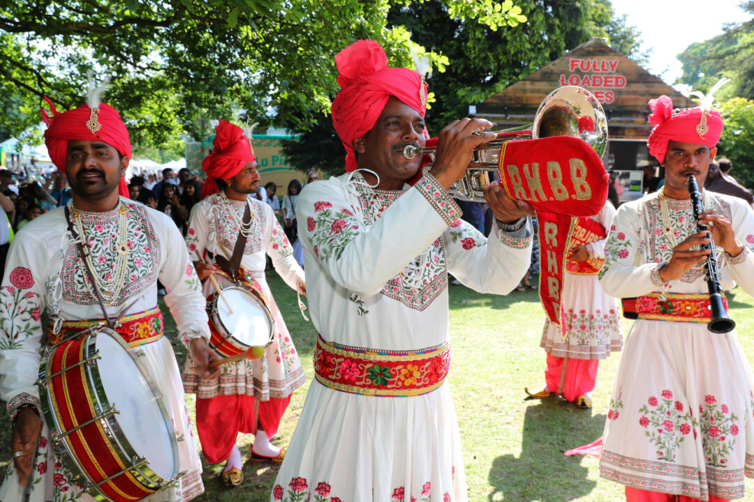 A lively outdoor performance featuring a traditional Indian brass band dressed in ornate white outfits with colourful embroidery and bright red turbans. Musicians play instruments including a trumpet, clarinet, and drums, with a red banner reading “RHBB” on the trumpet. The group stands on grass under trees, with festival stalls and people visible in the background on a sunny day.