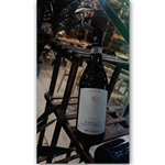 A bottle of 2014 Barolo wine labeled "Vinum Vita Est" and "Terre del Barolo" displayed on a wooden rack. The background features chairs and tables in an indoor dining setting, suggesting a restaurant atmosphere.