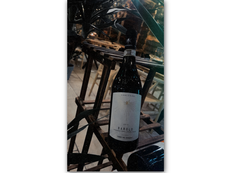 A bottle of 2014 Barolo wine labeled "Vinum Vita Est" and "Terre del Barolo" displayed on a wooden rack. The background features chairs and tables in an indoor dining setting, suggesting a restaurant atmosphere.