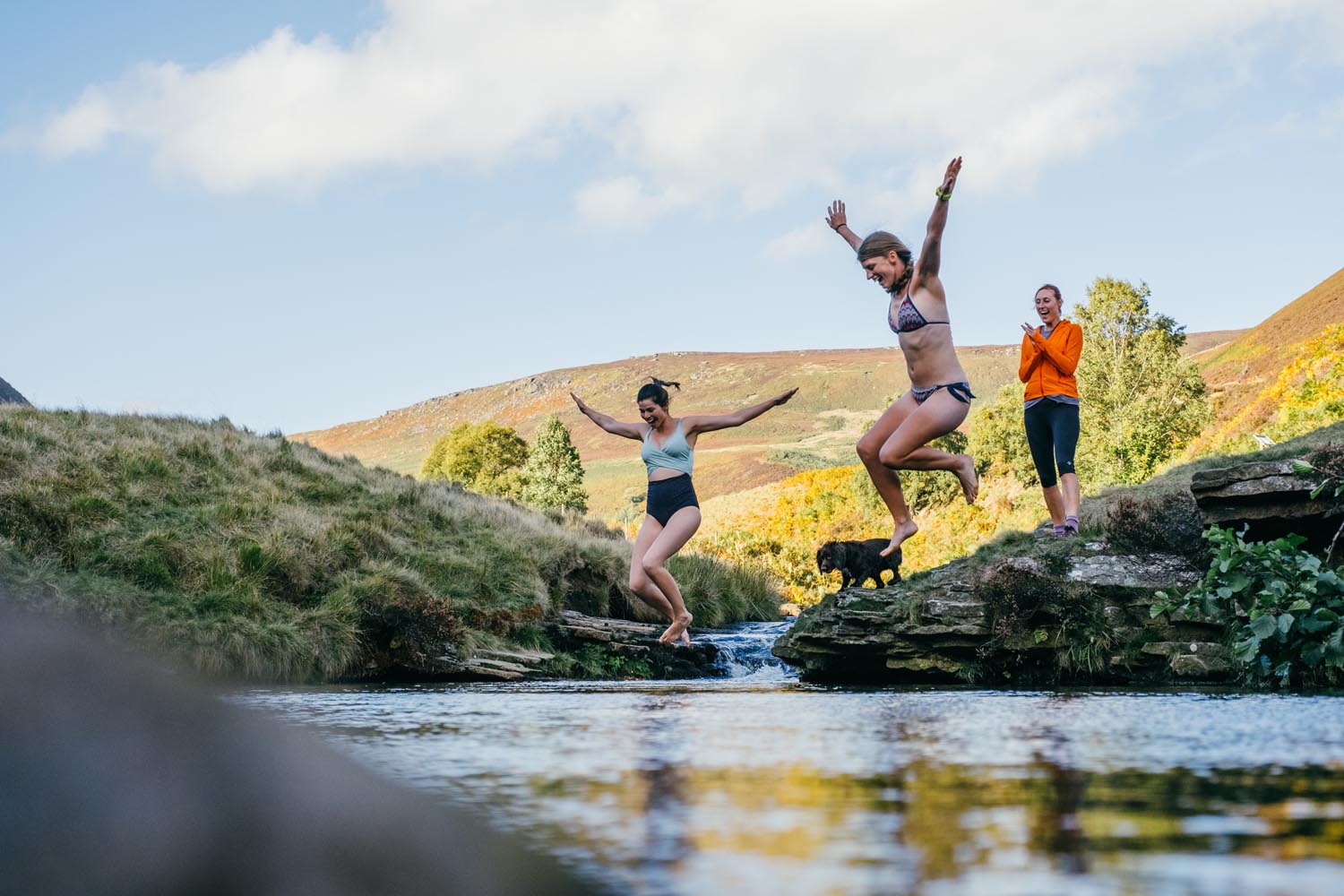 Three people wild swimming, two of them are jumping into a river while the third stands on the bank clapping.