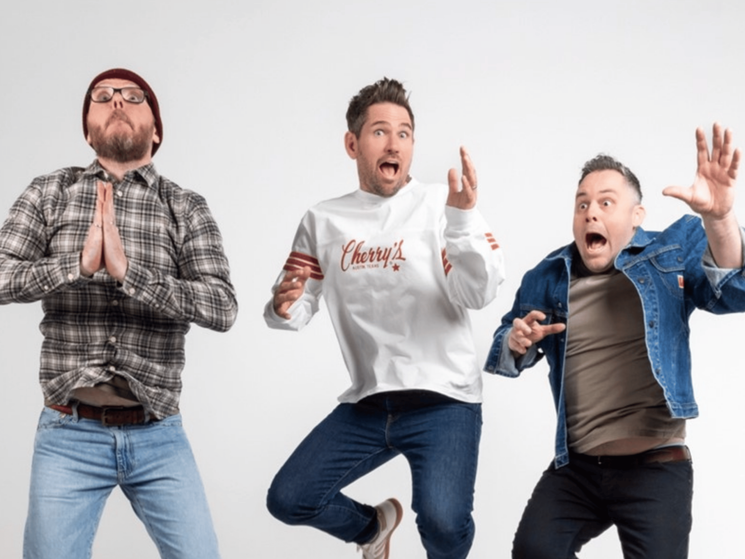 Three people are mid-jump against a plain white background. The person on the left wears a plaid shirt and jeans with hands pressed together in a prayer-like gesture. The person in the center wears a white sweatshirt with “Cherry’s” written on it and dark jeans, with arms bent and fingers pointing upward. The person on the right wears a denim jacket over a gray shirt and dark pants, with one arm raised and the other bent forward in an expressive pose.