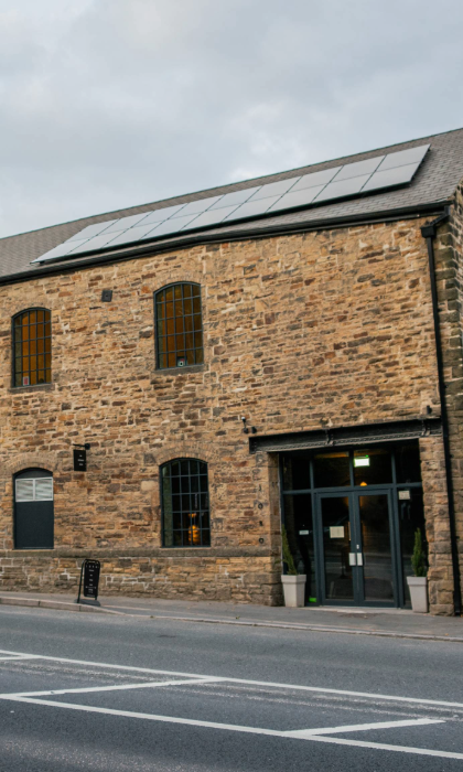 Exterior view of a large stone building with tall arched windows and a pitched roof fitted with solar panels. The structure has an industrial yet modern look, with a glass entrance door flanked by potted plants. It sits alongside a paved road with white markings, and the sky above is overcast. The building appears to be a converted mill or warehouse with contemporary features.