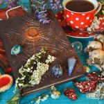 A vibrant tabletop scene shows two ornate, embossed journals decorated with crystals and small white flowers, arranged on a bright turquoise surface. Beside them is a red polka‑dot teacup and saucer filled with a dark hot drink, surrounded by dried herbs, a lit candle, and scattered decorative stones. Heart‑shaped biscuits with red icing and colourful dried flower petals complete the whimsical, cosy composition.