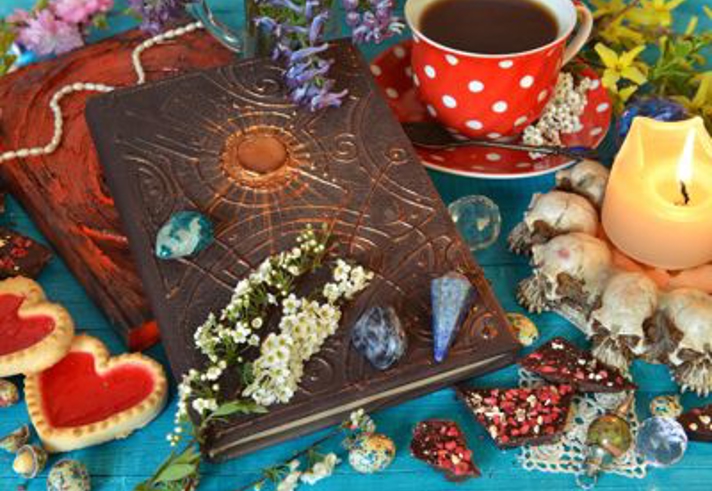 A vibrant tabletop scene shows two ornate, embossed journals decorated with crystals and small white flowers, arranged on a bright turquoise surface. Beside them is a red polka‑dot teacup and saucer filled with a dark hot drink, surrounded by dried herbs, a lit candle, and scattered decorative stones. Heart‑shaped biscuits with red icing and colourful dried flower petals complete the whimsical, cosy composition.