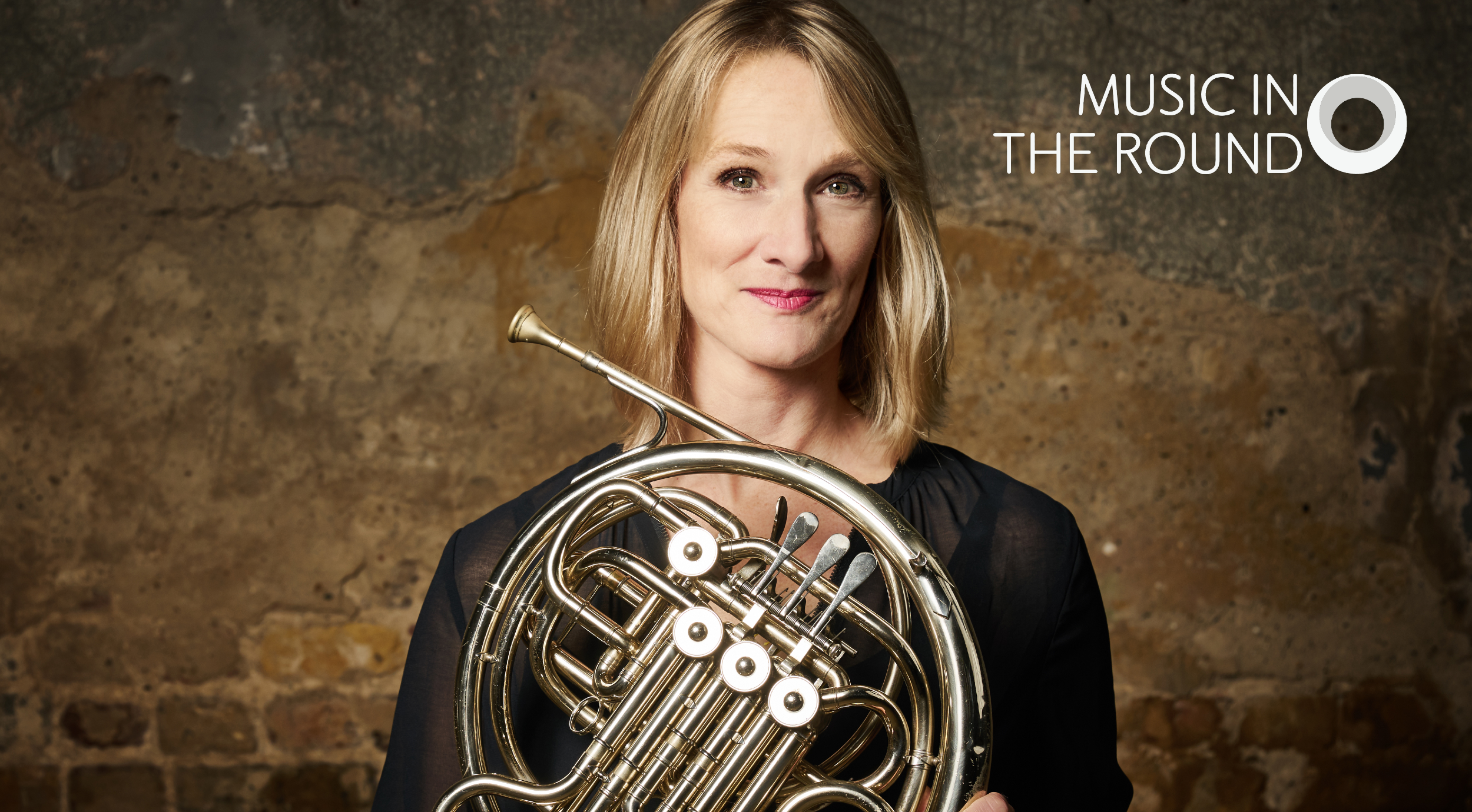 Person holding a French horn against a rustic stone wall background. The instrument’s shiny brass coils and valves are prominently displayed, reflecting light. In the top right corner, white text reads “Music in the Round” with a circular logo.