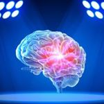 A glowing, semi‑transparent digital illustration of a human brain is shown floating at the center of a dark blue spotlighted stage. Bright white and blue lights arranged in circular clusters shine down from the upper left and upper right corners, illuminating the brain. The center of the brain glows with an intense red and pink light, highlighting internal structures against the deep blue background.
