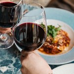Two wine glasses clinking above plates of food.
