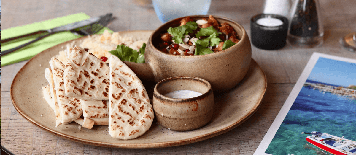 Food at Ego Mediterranean.  A rustic ceramic plate with folded grilled flatbreads, a small bowl of creamy white dip, and a larger bowl of stew topped with fresh herbs and sliced almonds. The plate sits on a wooden table alongside cutlery, a salt and pepper set, and a postcard featuring a coastal scene with turquoise water and a red-and-white boat.