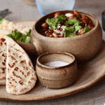 Food at Ego Mediterranean.  A rustic ceramic plate with folded grilled flatbreads, a small bowl of creamy white dip, and a larger bowl of stew topped with fresh herbs and sliced almonds. The plate sits on a wooden table alongside cutlery, a salt and pepper set, and a postcard featuring a coastal scene with turquoise water and a red-and-white boat.