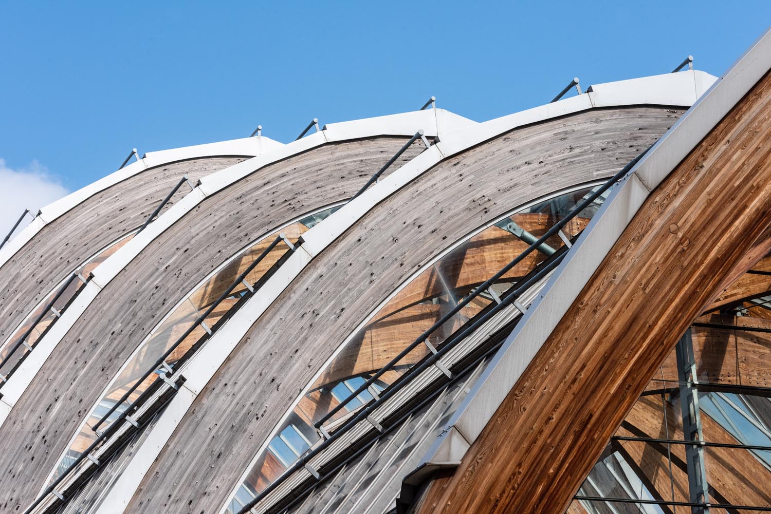 Close-up view of large curved wooden arches forming part of a modern architectural structure. The arches are supported by metal framework and glass panels, with reflections of surrounding buildings visible. The design features repeating geometric patterns against a clear blue sky.