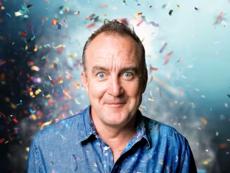 A portrait of Jimeoin. Behind him there is loads of confetti flying about.
