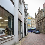 A narrow cobbled street lined with older buildings, with a large shop window in the foreground displaying a sign that reads “7THMAGPIE.” Parked cars and a tram are visible further down the street.