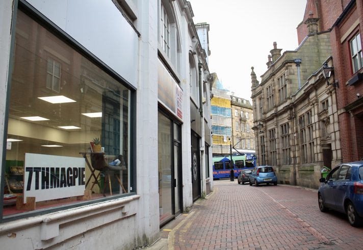 A narrow cobbled street lined with older buildings, with a large shop window in the foreground displaying a sign that reads “7THMAGPIE.” Parked cars and a tram are visible further down the street.
