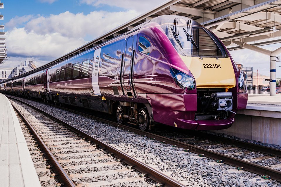 Modern maroon and cream-colored train with the number 222104 parked at a station platform under a partially covered roof. The train has a sleek, aerodynamic design with reflective windows, and the tracks are lined with gravel. The sky is partly cloudy, and station signage is visible in the background.