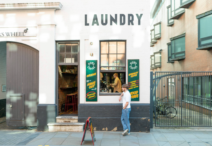 Exterior of a white-painted building with the word “LAUNDRY” in bold black letters above two windows and a doorway. The entrance is open, revealing a rustic interior with bar stools and a counter. Green signs with yellow text advertise coffee and good vibes. A person stands outside on the pavement near a sandwich board, while another is visible inside by the counter. The setting is urban, with a gated area and bicycles to the right.