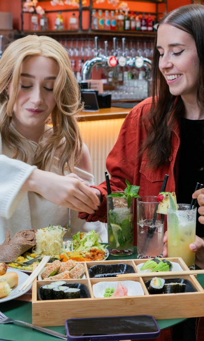 Close-up of a green table in a lively indoor food hall, filled with assorted dishes and drinks. A wooden bento-style tray holds sushi rolls, nigiri, sesame-coated pieces, and pickled ginger with chopsticks resting on top. Nearby plates feature flatbread, shredded cabbage, and colourful vegetable sides. Three tall cocktails garnished with fruit and herbs are also visible, with a bar lined with bottles in the background.
