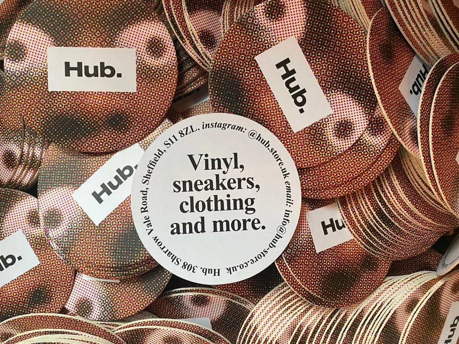 A close-up of a pile of Hub. merchandise stickers. In the middle is a white circle with the words 'VINYL, SNEAKERS, CLOTHING AND MORE'.