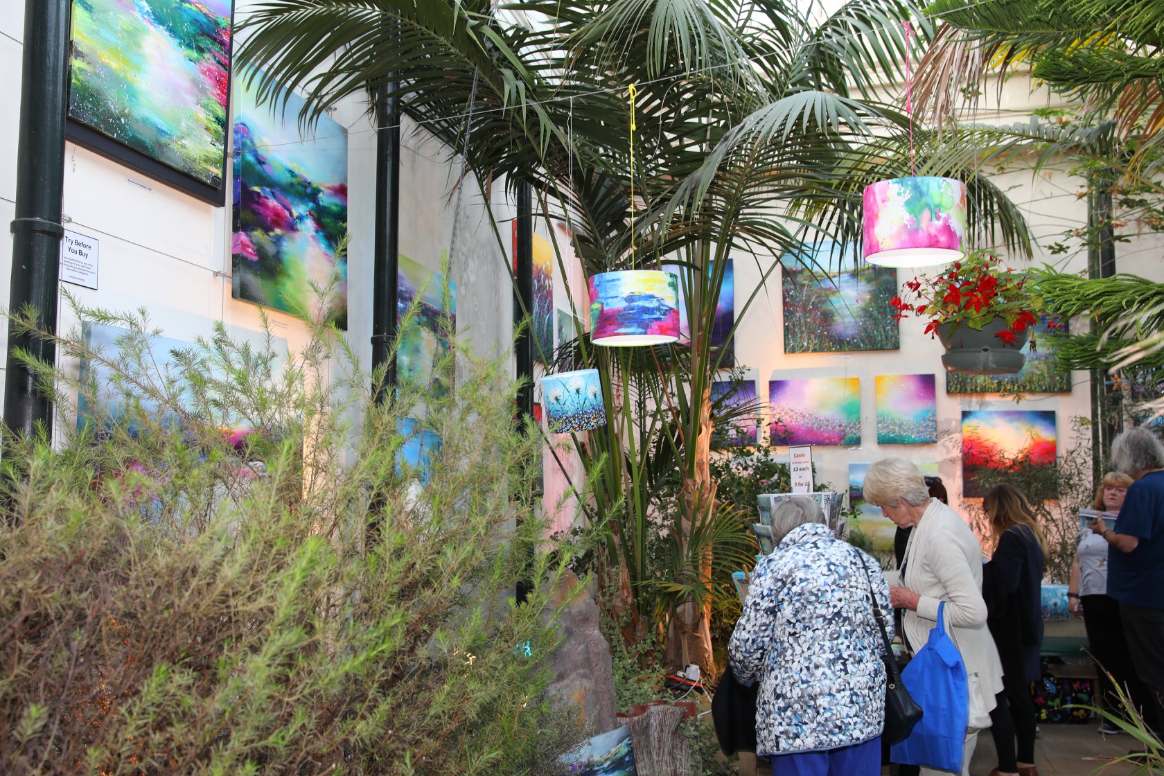 An indoor art display features vibrant abstract paintings in bold colours mounted on cream walls, with several matching lampshades hanging among tall green palm plants. A group of people are viewing the artwork and browsing items on tables. The scene is bright and colourful, with a mix of natural greenery and artistic decor creating a lively exhibition atmosphere.