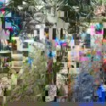 An indoor art display features vibrant abstract paintings in bold colours mounted on cream walls, with several matching lampshades hanging among tall green palm plants. A group of people are viewing the artwork and browsing items on tables. The scene is bright and colourful, with a mix of natural greenery and artistic decor creating a lively exhibition atmosphere.