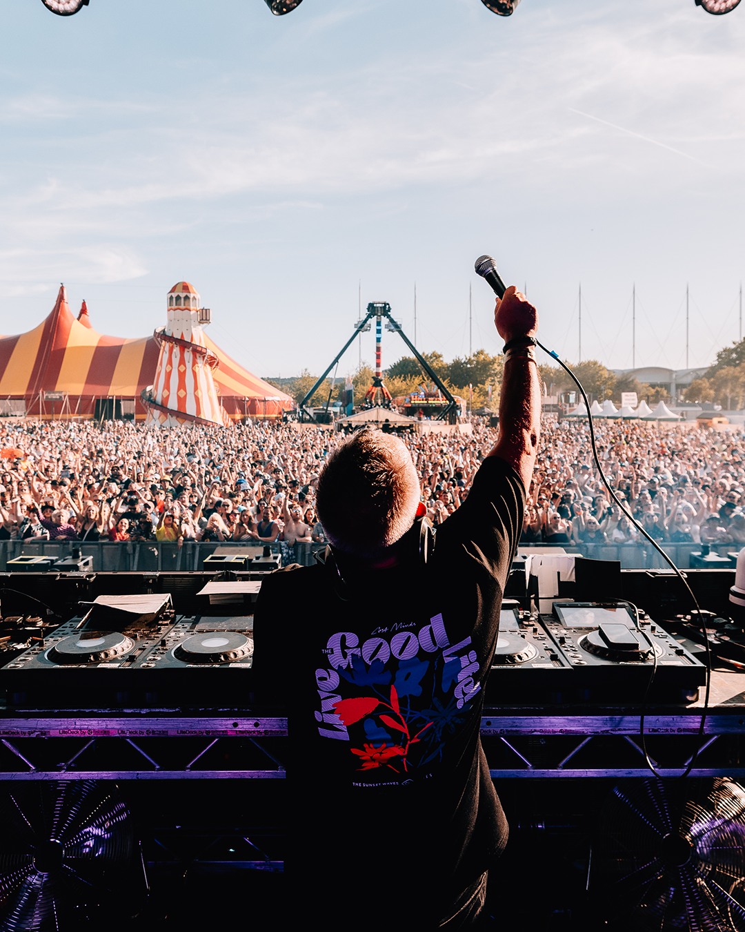 A DJ stands behind a mixing console on an outdoor stage, holding a microphone up toward a large crowd. The audience stretches far into the distance, with a colorful striped circus-style tent and a tall amusement ride visible in the background under a clear blue sky.