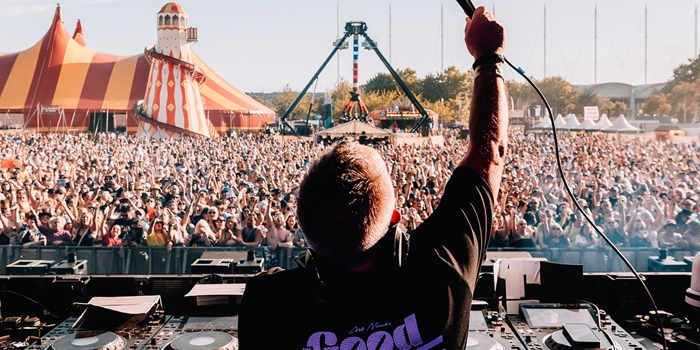 A DJ stands behind a mixing console on an outdoor stage, holding a microphone up toward a large crowd. The audience stretches far into the distance, with a colorful striped circus-style tent and a tall amusement ride visible in the background under a clear blue sky.