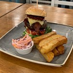 A tall burger , a stack of chunky square cut chips and a bowl of coleslaw on a square plate.