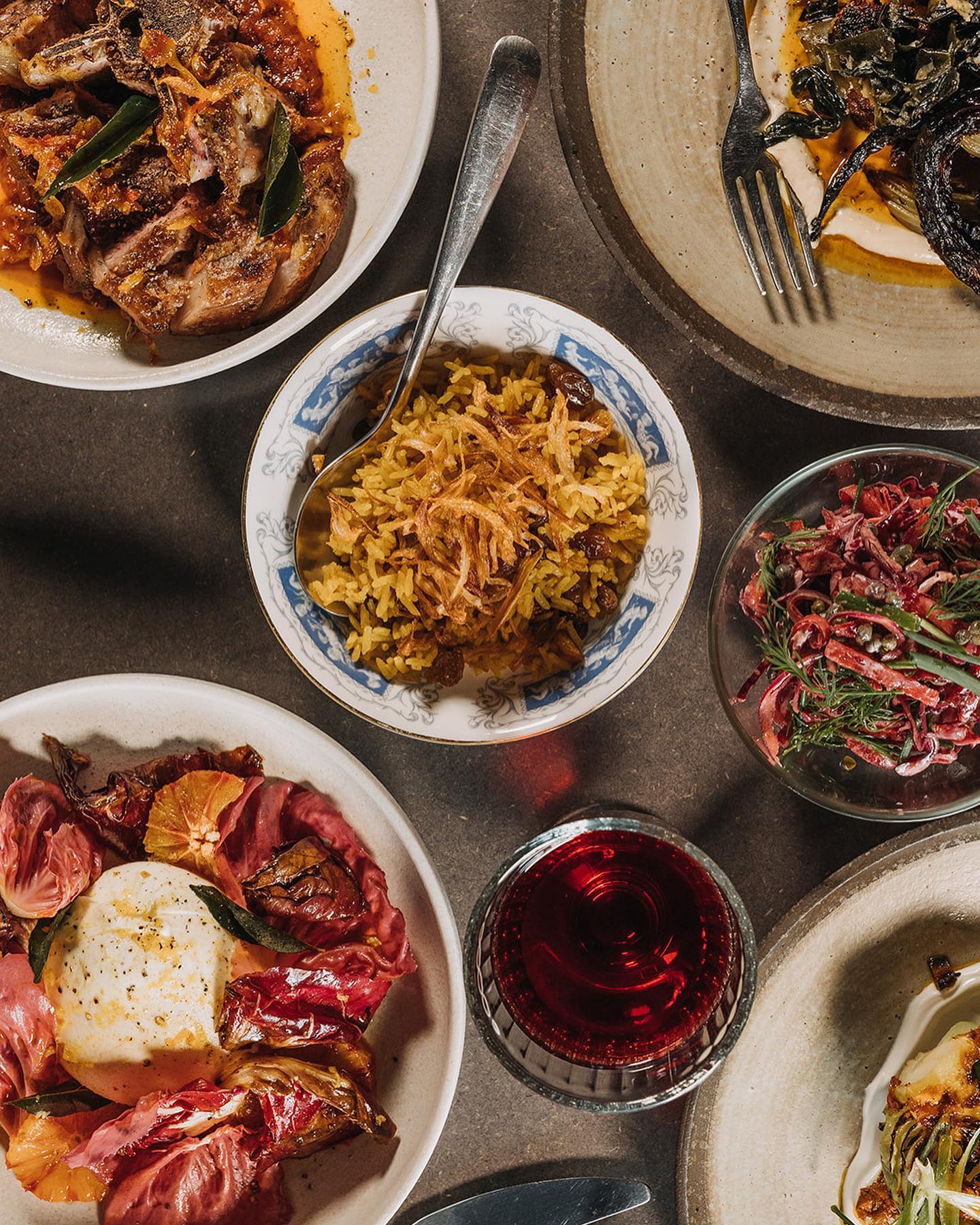 A tabletop spread of vibrant dishes including grilled meat in sauce, a bowl of spiced rice topped with crispy onions, a salad with red leaves and herbs, and a plate with creamy cheese and roasted vegetables, alongside a glass of red drink.