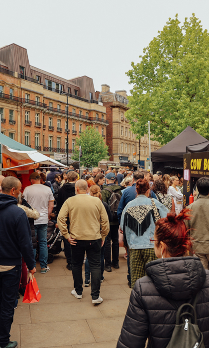 Crowds walking through an outdoor market lined with food stalls and tents in a city square