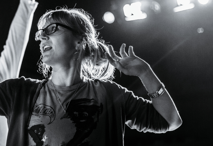 A black and white photo standing under stage lights, with one arm slightly raised. Another person's arm is just coming into shot.