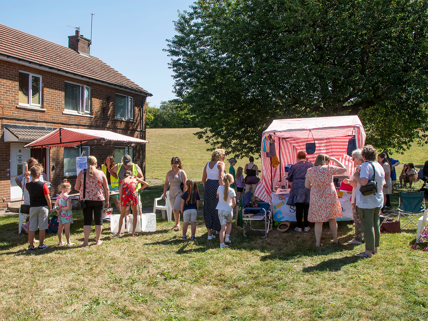 People gathered outdoors at a summer fair on a grassy area near a brick house. A red-and-white striped stall is set up under a large tree, with several individuals standing around it. Others are chatting or waiting nearby, and some children are present. Folding chairs and a small canopy are visible, suggesting a casual community event in warm weather.