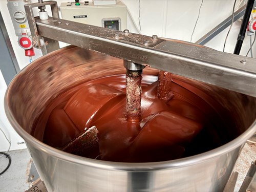 Large stainless steel mixing machine filled with thick, glossy melted chocolate being stirred by rotating metal paddles. Control panels and safety equipment are visible in the background.