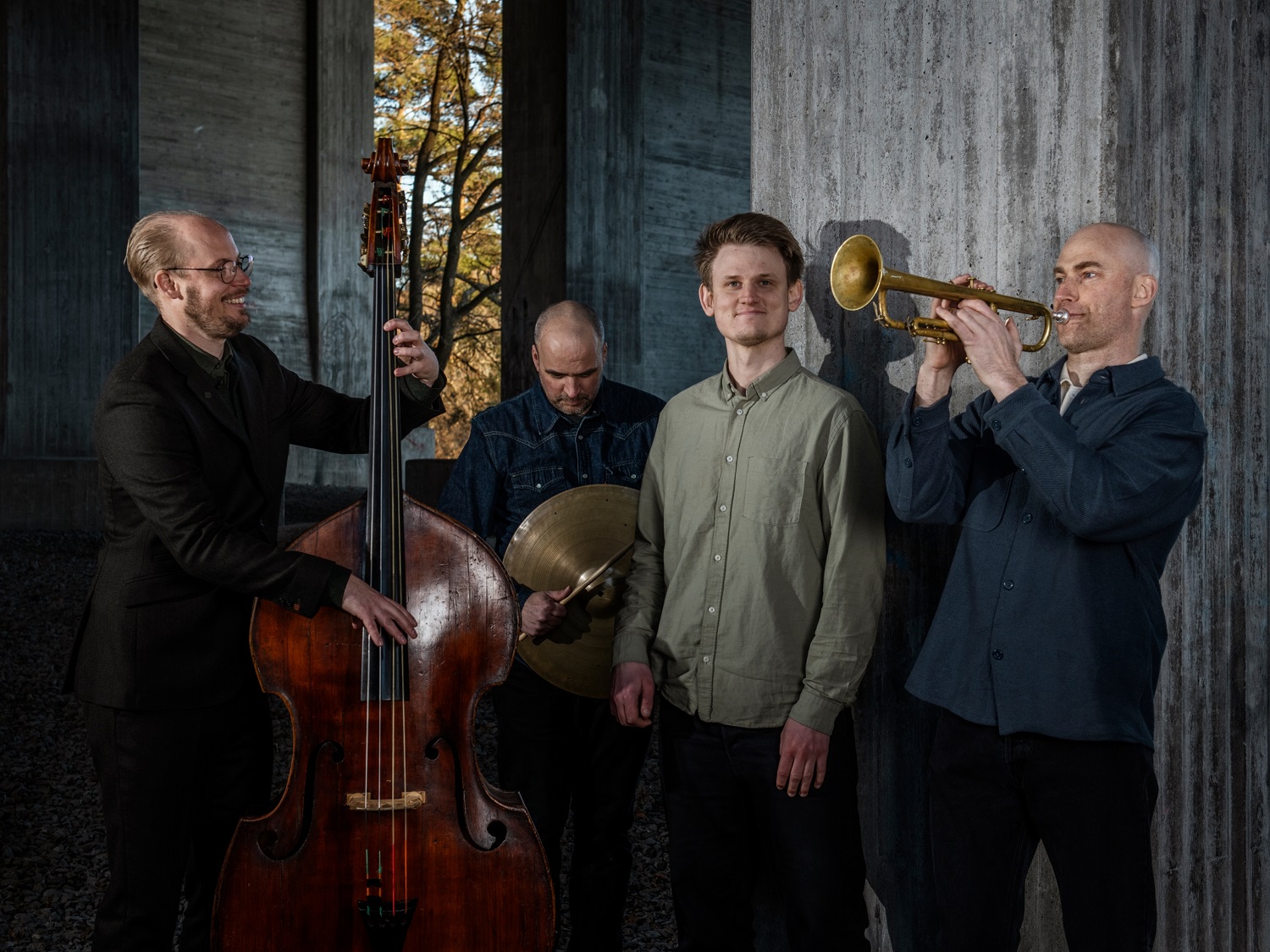 A four‑piece jazz group stands in a concrete architectural space. One musician plays a double bass, another holds cymbals, a third stands in the centre, and the fourth plays a trumpet. Sunlight filters through an opening behind them, creating contrast between the warm outdoor light and the cool, shadowed interior.