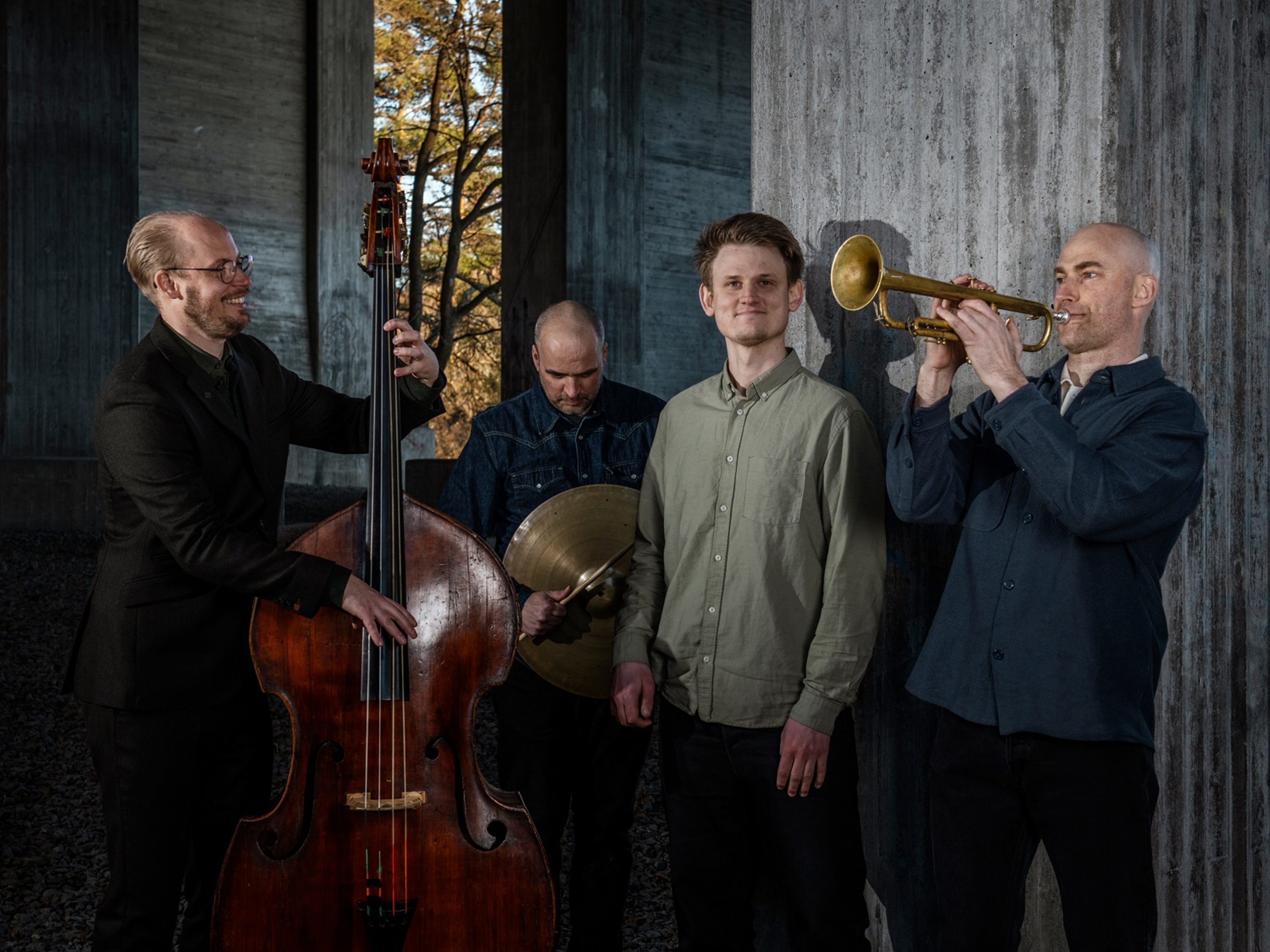 A four‑piece jazz group stands in a concrete architectural space. One musician plays a double bass, another holds cymbals, a third stands in the centre, and the fourth plays a trumpet. Sunlight filters through an opening behind them, creating contrast between the warm outdoor light and the cool, shadowed interior.