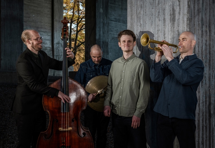 A four‑piece jazz group stands in a concrete architectural space. One musician plays a double bass, another holds cymbals, a third stands in the centre, and the fourth plays a trumpet. Sunlight filters through an opening behind them, creating contrast between the warm outdoor light and the cool, shadowed interior.