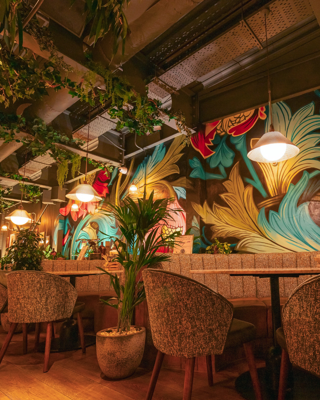 A restaurant interior featuring bold, colourful botanical wall art, hanging plants, cushioned seating, and warm pendant lighting.